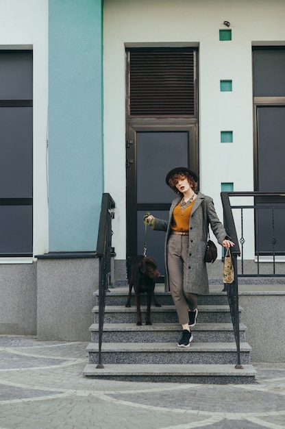 Signora carina in vestiti alla moda e cappello pone sulla fotocamera con un cucciolo al guinzaglio