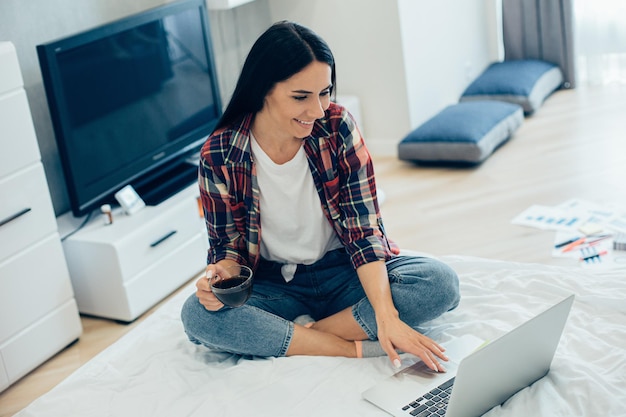 Signora caucasica allegra che si siede con una tazza di caffè sul suo letto e che utilizza un computer portatile moderno