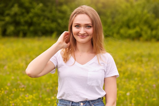 Signora con i capelli biondi e gli occhi azzurri, sembra romantica e fresca, posa in prato, toccandosi i capelli, indossando maglietta bianca casual, trascorrendo giornate estive nella natura con il suo fidanzato. Concetto di stile di vita.