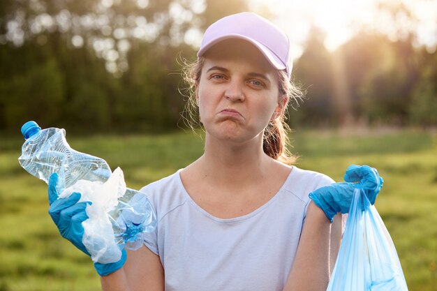 Signora con la faccia contorta che indossa guanti di lattice blu, con in mano la spazzatura, con espressione facciale turbata, vuole ripulire il pianeta dai rifiuti e riutilizzare i rifiuti, problemi ecologici.