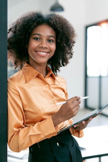 Signora d'affari afroamericana positiva utilizzando la tavoletta digitale in piedi in ufficio sorridendo alla telecamera