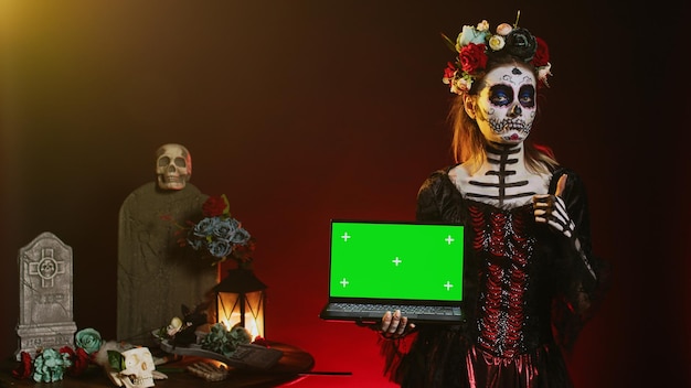 Signora dei morti che tiene in mano un laptop con schermo verde, che mostra il display copyspace isolato sul PC. Modello di Halloween con body art inquietante che ha isolato il modello di mockup chroma key in studio.