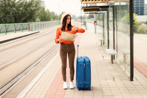 Signora del viaggiatore preoccupata con la valigia che controlla l'orologio alla stazione ferroviaria