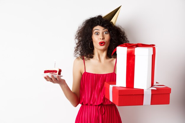 Signora divertente in abito rosso e cappello da festa, festeggia il compleanno, tiene regali di b-day e torta con la candela, guardando divertita alla telecamera, in piedi su sfondo bianco.