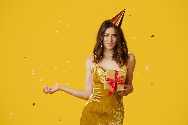 Signora felice che tiene confezioni regalo avvolte indossando abito festivo e cappello di compleanno sfondo giallo con coriandoli che cadono