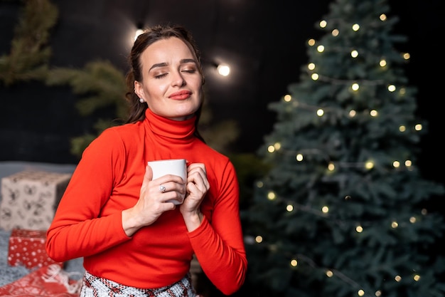 Signora in maglione rosso con in mano una tazza bianca Ragazza sognante con gli occhi chiusi vicino all'albero di Natale Donna con una tazza di tè circondata da regali e albero di Natale