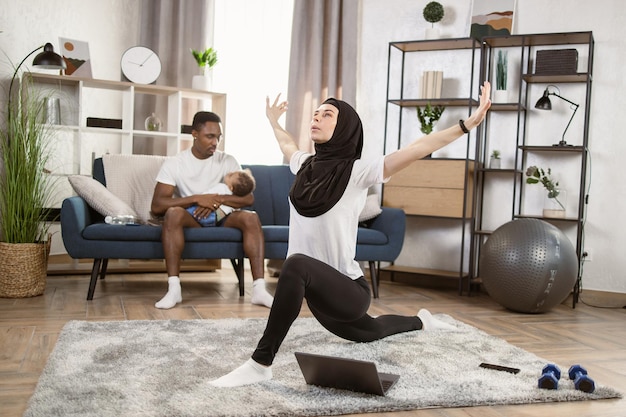 Signora musulmana In hijab che fa allenamento pilates con il computer portatile a casa praticando yoga online