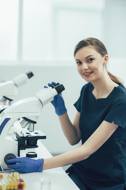 Signora positiva che lavora in un laboratorio sterile e sorride mentre è seduta con un microscopio