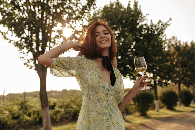 Signora positiva con l'acconciatura allo zenzero e la benda sul collo in abiti verdi e cappello che sorride e tiene con champagne all'aperto