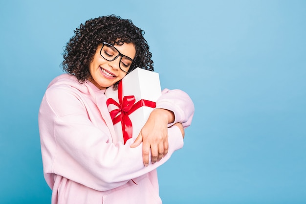 Signora riccia afroamericana felice nella risata casuale mentre tiene il contenitore di regalo presente isolato sopra fondo blu.