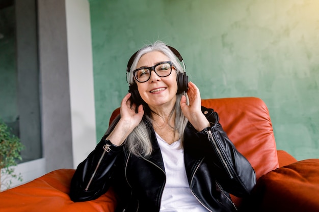 Signora senior felice allegra affascinante con lunghi capelli grigi, indossa elegante giacca di pelle nera, seduto in poltrona rossa su sfondo verde