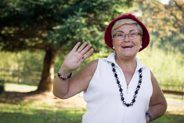 Signora sorridente anziana con cappello rosso e occhiali che salutano in una giornata di sole nel parco