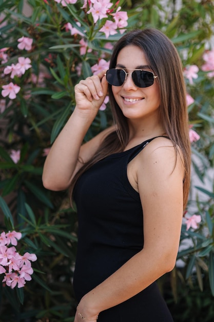 Signora sorridente in abito nero in piedi davanti a fiori donna in occhiali da sole in posa per il fotografo