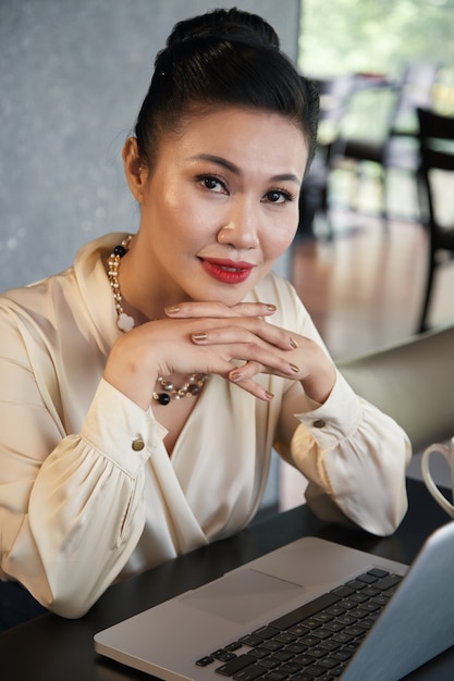 Signora sorridente positiva di affari che si siede alla tavola del ristorante con il computer portatile aperto e che esamina macchina fotografica