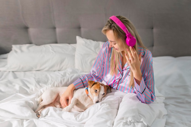Signora spensierata in pigiama che ascolta musica in cuffia mentre è seduta a letto con il suo amato animale domestico
