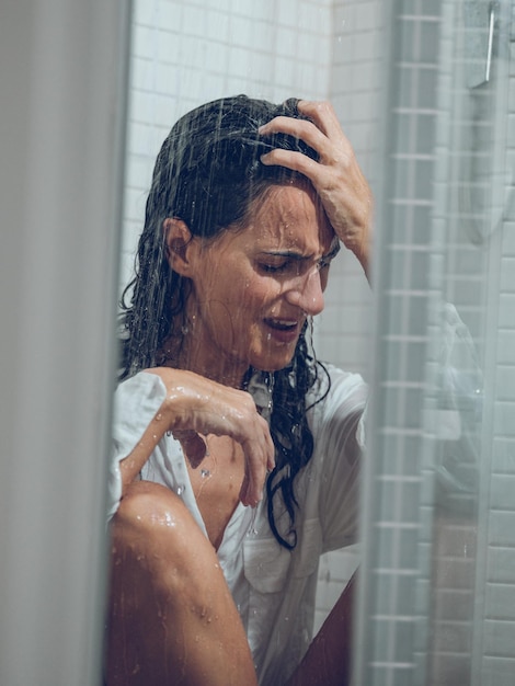 Signora vestita dai capelli lunghi che piange nella cabina della doccia mentre è seduta sotto gli spruzzi d'acqua e si tocca la fronte