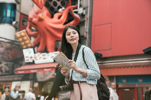 signora viaggiatore che tiene mappa in piedi sulla strada trafficata del dotonbori. la fotografa femminile viaggia ad osaka. felice turista femminile che cerca la strada per l'hotel in una città straniera durante le vacanze.