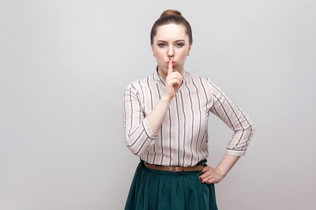 Silenzio! Bella giovane donna seria in camicia a righe e gonna verde con trucco e acconciatura di divieto raccolto, in piedi con il dito sul naso e segno silenzioso. girato in studio, isolato su sfondo grigio