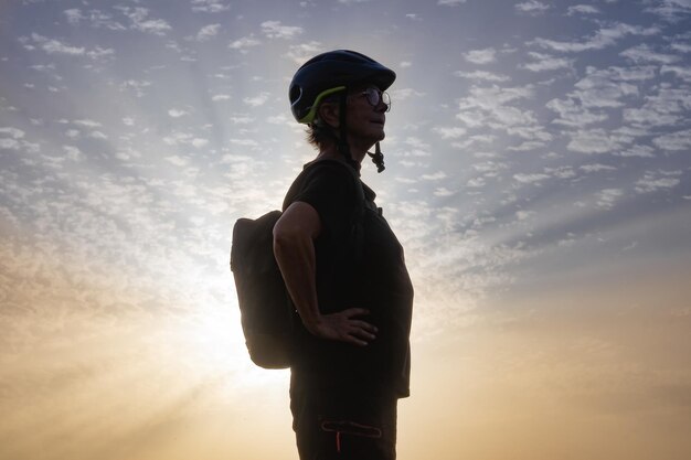 Silhouette di donna attiva senior che indossa casco da ciclismo e zaino in piedi con una bellissima luce del tramonto sullo sfondo