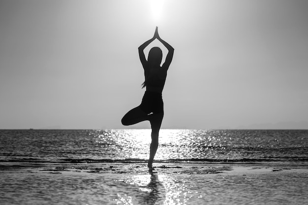 Silhouette di donna in piedi in posa yoga sulla spiaggia tropicale durante il tramonto Ragazza caucasica che pratica yoga vicino all'acqua di mare In bianco e nero