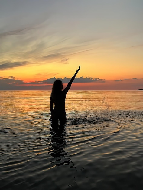 silhouette di giovane donna al tramonto arancione nell'onda di acqua di mare e cielo nuvoloso rosa