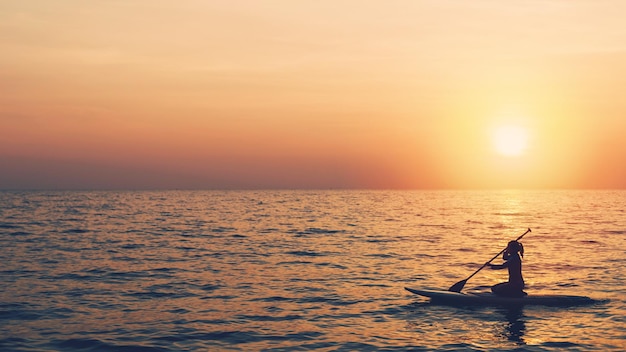 Silhouette di giovane donna che gioca paddle board in mare con lo sfondo del cielo al tramonto. Concetto di sport acquatici e viaggi in estate.