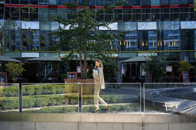 Silhouette di giovane donna d'affari in abito beige che cammina nel centro della città in posa vicino a edifici per uffici