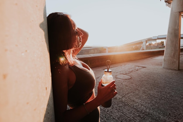 Silhouette di giovane donna in appoggio contro un muro con in mano una bottiglia d'acqua durante un tramonto colorato con spazio per la copia Riposo dopo l'allenamento su strada urbana concetto ambientale Ragazza marocchina