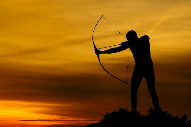 Silhouette di un bell'uomo con un'antica arma arco e freccia su uno sfondo di cielo e tramonto