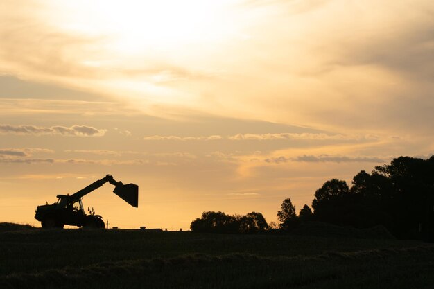 Silhouette di un escavatore funzionante con una benna rialzata sullo sfondo del tramonto e delle nuvole Il campo è gestito da macchine agricole La stagione della raccolta o della semina
