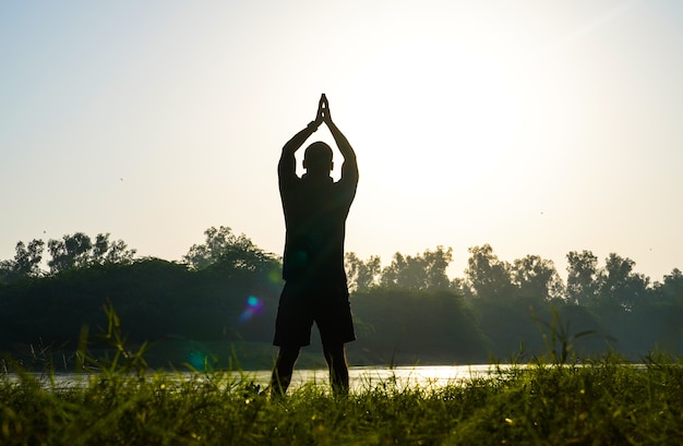 Silhouette di un ragazzo che fa surya namaskar nel parco vicino al sole e al fiume - concetto di salute