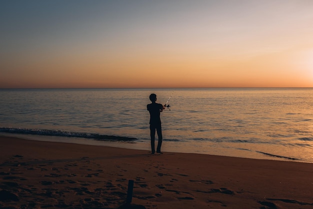Silhouette di un uomo che pesca sullo sfondo di un bellissimo tramonto colorato accanto al mare piacevole sulla spiaggia sabbiosa.