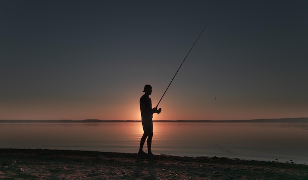 Silhouette di un uomo con una canna da pesca sulla riva del lago sullo sfondo del cielo al tramonto