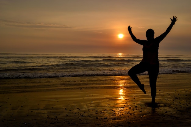 Silhouette di una donna con le braccia aperte e su un piede sulla riva di una spiaggia al tramonto