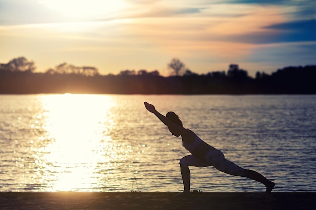 Silhouette di una giovane donna che fa esercizi di yoga sulla spiaggia del lago al tramonto