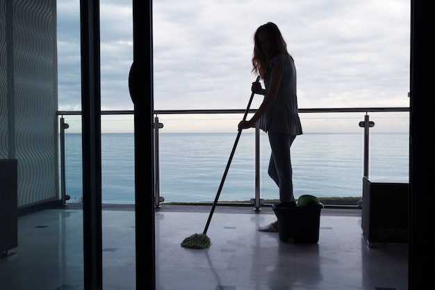 silhouette di una giovane donna che lava una scopa con un pavimento di marmo su un balcone all'aperto