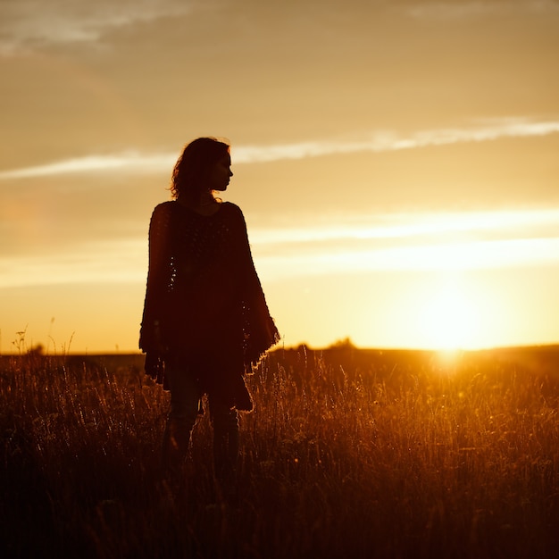 Silhouette di una giovane donna felice al tramonto, ragazza all'aperto in un poncho scozzese in un campo con spighette