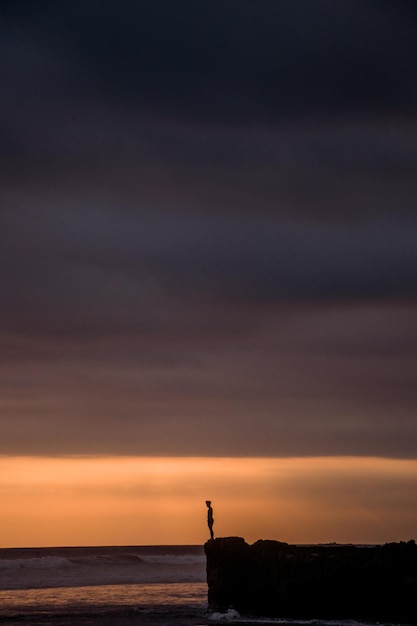 Silhouette di una persona in piedi su una scogliera contro il mare durante il tramonto