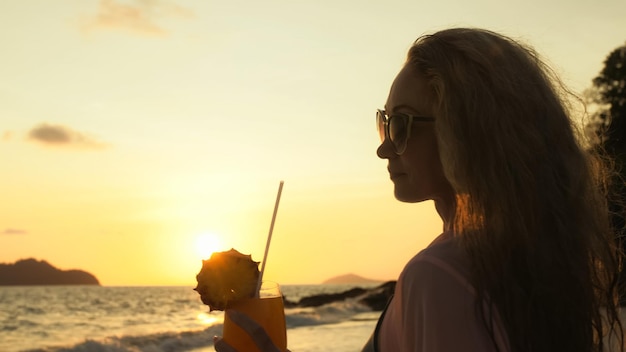 Silhouette donna cammina sul tramonto dorato sulla spiaggia tropicale sullo sfondo del mare Ragazza in tunica rosa mantello camicia di seta beve il suo cocktail arancione Pina Colada con fetta di ananas