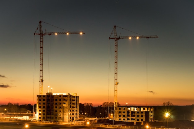 Silhouette gru a torre e edifici alti multipiano incompiuti in costruzione al tramonto nel deserto su un cantiere illuminato