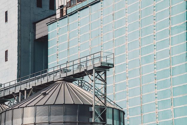 Silos agricoli. Conservazione e essiccazione di cereali, grano, mais, soia, girasole. Esterno di un edificio industriale. Primo piano di grandi contenitori metallici d'argento. Sfondo di carri armati agricoli con spazio di copia.