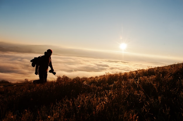 Siluetta del fotografo dell'uomo con le montagne del fondo della macchina fotografica a disposizione sul tramonto con nebbia