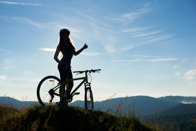 Siluetta del motociclista femminile felice con la bicicletta nelle montagne, indossando casco, mostrando i pollici sulla mattina di estate contro il cielo blu. Attività sportiva all'aperto, concetto di lifestyle. Copia spazio