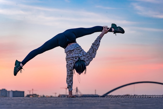 Siluetta dell'artista di circo femminile flessibile che fa verticale sul tramonto drammatico e sul paesaggio urbano. Concetto di individualità, creatività ed eccezionale.