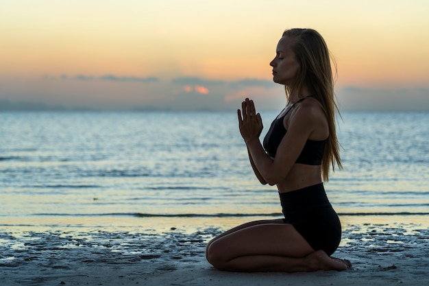Siluetta della donna che si siede alla posa di yoga sulla spiaggia tropicale durante il tramonto. Ragazza caucasica che pratica yoga vicino all'acqua di mare.