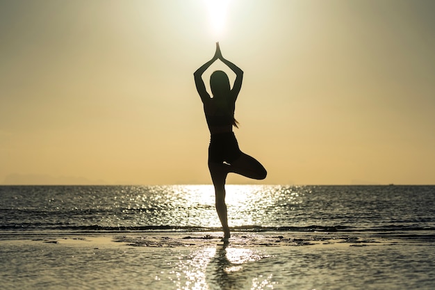 Siluetta della donna che sta alla posa di yoga sulla spiaggia tropicale durante il tramonto. Ragazza caucasica che pratica yoga vicino all'acqua di mare