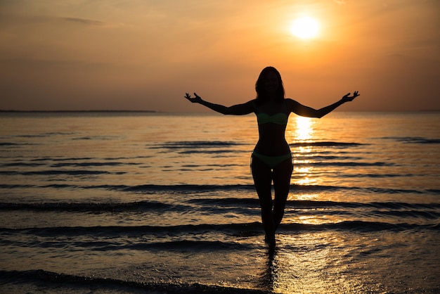 Siluetta della donna felice in bikini che posa sulla spiaggia al tramonto
