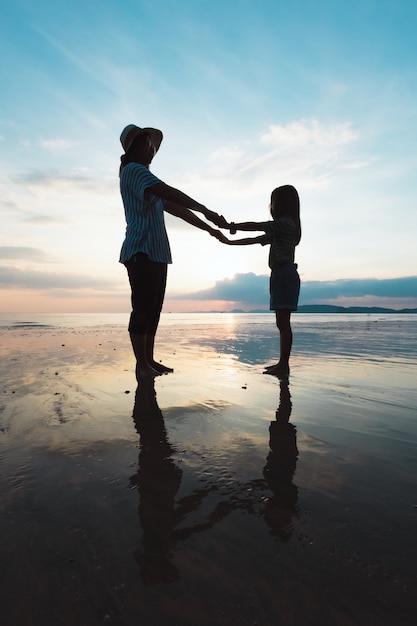 Siluetta della madre asiatica e della figlia che tengono la mano e che giocano insieme sulla spiaggia all'ora del tramonto con il bello mare e cielo. La famiglia gode con il concetto di natura.