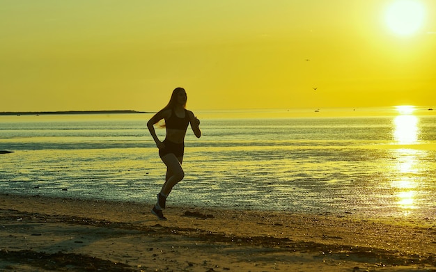 Siluetta della ragazza che corre in riva al mare sulla spiaggia all'alba, sfondo spiaggia nera al tramonto con il sole. Attività sportiva di jogging fitness donna in estate