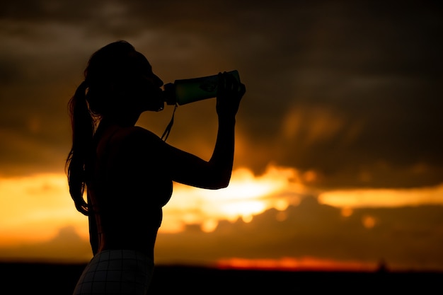 Siluetta della ragazza in abiti sportivi beve l'acqua da una bottiglia al tramonto.